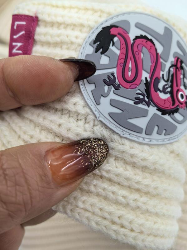 Close-up of cream chunky knit winter beanie hat held by hand with glitter ombre nails, featuring round pink snake rubber patch badge