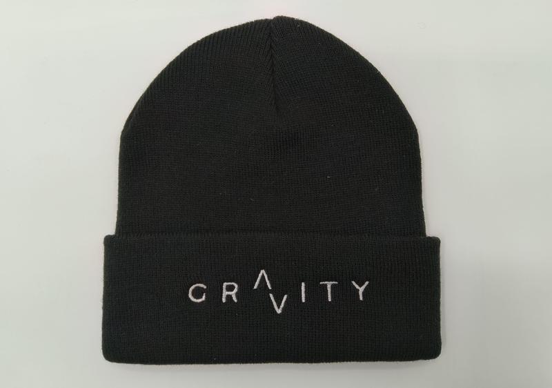 Black ribbed knit cuffed beanie hat with white embroidered GRAVITY logo on folded cuff, laid flat on white background