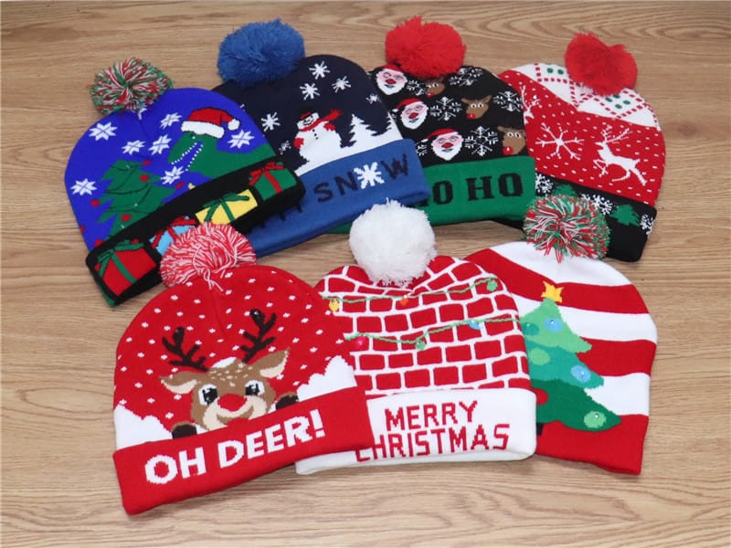 Six colorful Christmas knit beanies with pom-poms featuring reindeer, Santa, snowman, and holiday tree patterns on wooden surface
