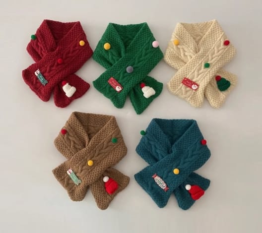 Five colorful children's cable knit crossover scarves in red, green, cream, brown, and teal with festive pom-pom and holiday charm decorations