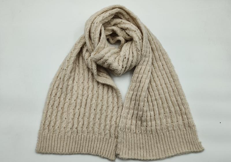 Cream beige cable knit scarf loosely knotted on white background, showing textured aran pattern and ribbed hem edges