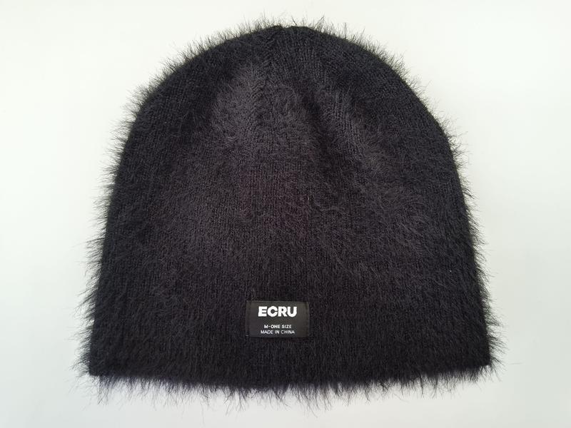 Black fluffy knit beanie hat with furry texture and small label patch, laid flat on white background