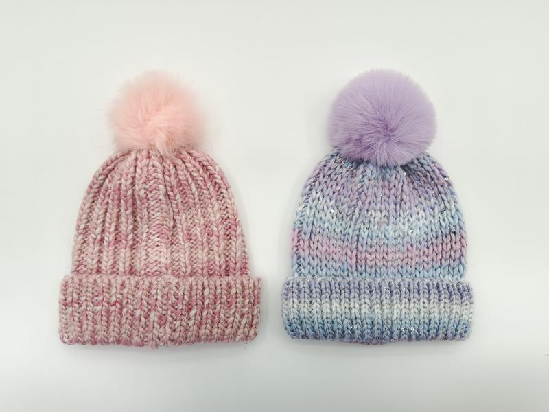 Two chunky knit pom-pom beanies side by side: pink marl and lavender blue tie-dye with faux fur pom-poms on white background