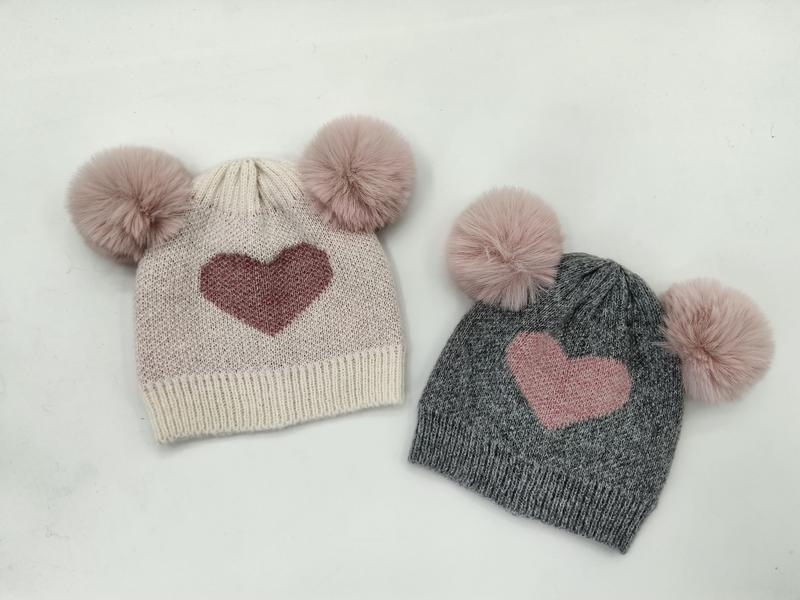 Two kids knit beanies with double faux fur pom poms and heart motif, one cream and one gray