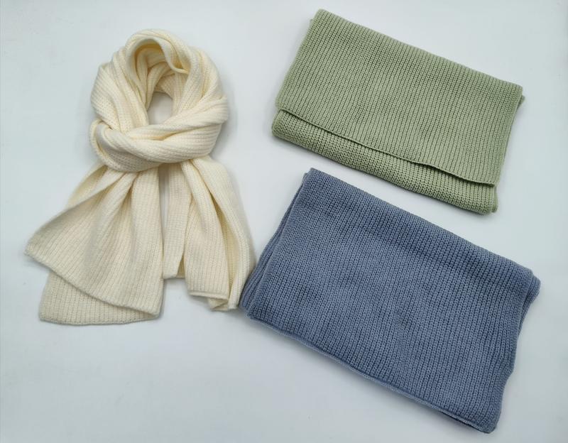 Three ribbed knit winter accessories on white background: cream scarf loosely tied, folded sage green and steel blue neck warmers