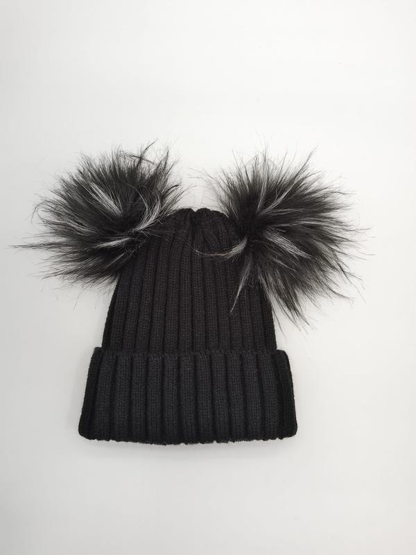 Black ribbed knit winter beanie hat with two large faux fur pom poms on top, laid flat on white background