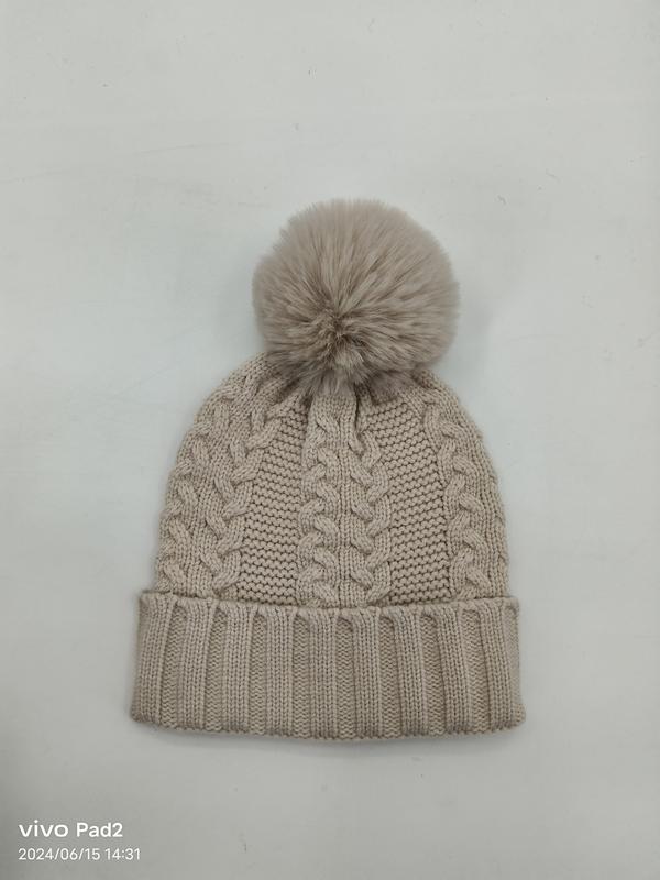 Beige cable knit winter beanie hat with large faux fur pom pom and ribbed cuff, laid flat on white background
