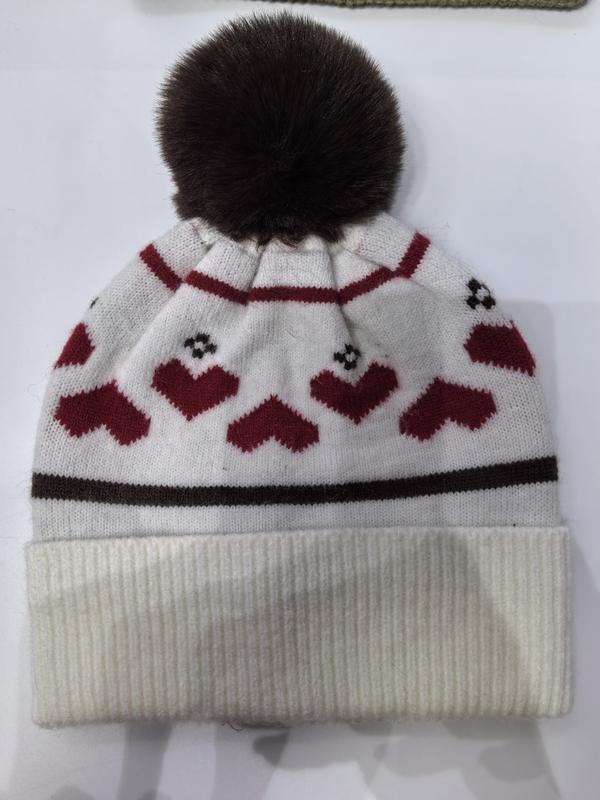 White knitted winter beanie hat with red heart pattern, dark brown faux fur pompom, and ribbed cuff on white background