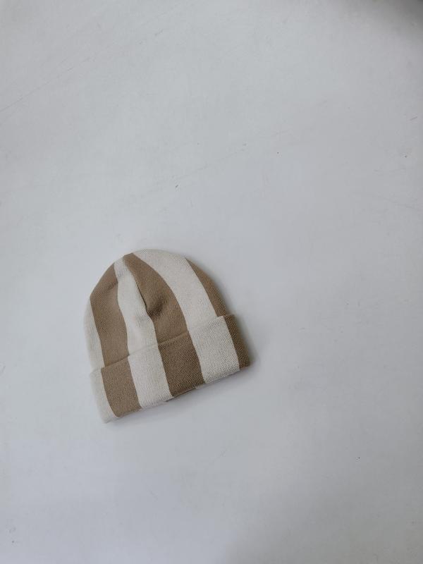 Small knitted beanie hat with wide tan and cream vertical stripes laid flat on white surface