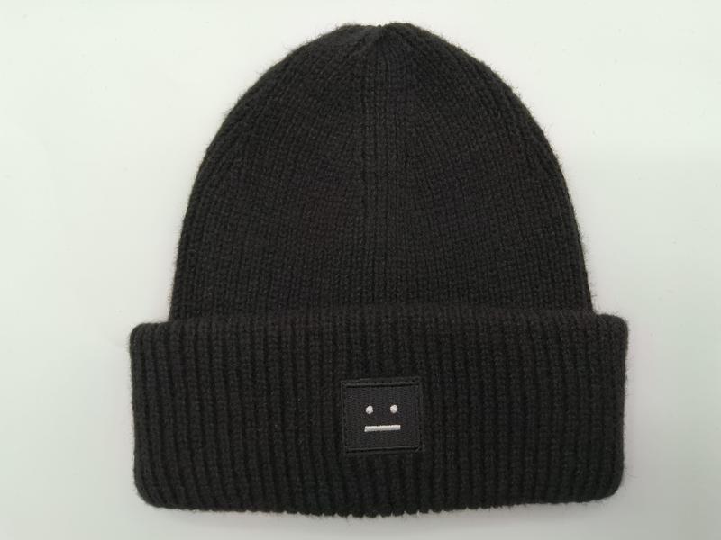 Black ribbed knit beanie hat with folded cuff and small embroidered face patch logo on white background