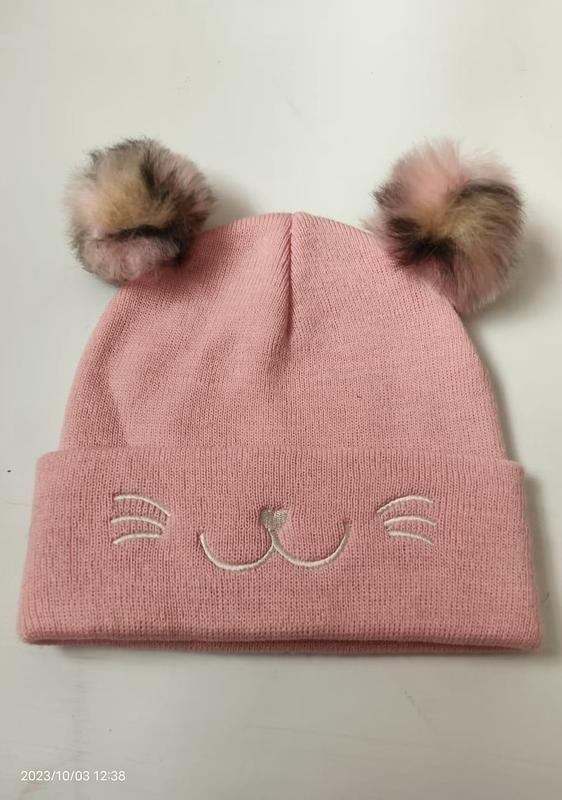 Pink knitted winter beanie hat with two faux fur pom poms and embroidered cat whiskers and nose on the cuff